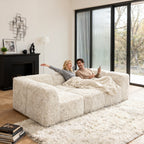 Soft sand daybed –showcased in a modern cozy living room setting. A model sits on it to demonstrate scale and comfort